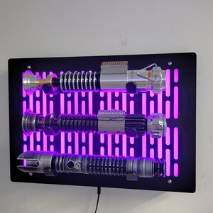 May include: A black wall mount with a purple light display and three silver lightsaber replicas mounted on it.