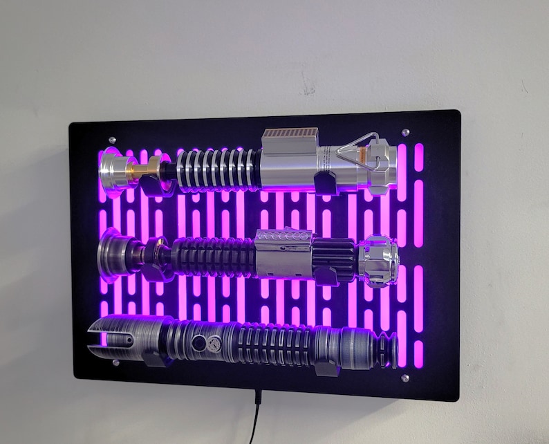 May include: A black wall mount with a purple light display and three silver lightsaber replicas mounted on it.