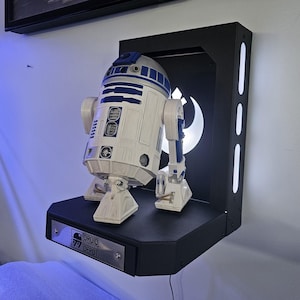 May include: A black wall shelf with a white and blue R2-D2 robot figure on a black platform. The shelf has a white light behind the robot and a black plaque with the text "Droid 77 Depot".