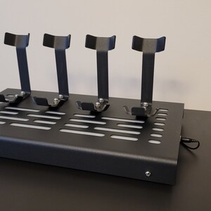 Pode incluir: Um suporte de metal preto com tr&ecirc;s suportes verticais para segurar fones de ouvido. O suporte tem uma base perfurada e um cabo preto.