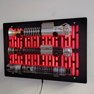 May include: A black wall mount with a red light grid pattern, displaying three silver and black lightsabers. The lightsabers are mounted on the wall with a black metal bracket.