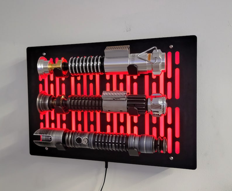 May include: A black wall mount with a red light grid pattern, displaying three silver and black lightsabers. The lightsabers are mounted on the wall with a black metal bracket.