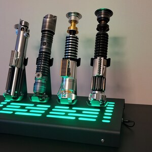 Pode incluir: Quatro r&eacute;plicas de sabres de luz em um suporte preto com uma luz verde. Os sabres de luz s&atilde;o prateados, pretos, dourados e verdes.
