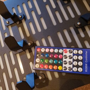 May include: A black and white grid panel with multiple black plastic hooks and a remote control for RGBW lights. The remote control has a variety of color options and settings, including 25%, 50%, 75%, and 100% brightness, jump, fade, flash, auto, and slow.