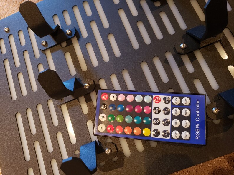 May include: A black and white grid panel with multiple black plastic hooks and a remote control for RGBW lights. The remote control has a variety of color options and settings, including 25%, 50%, 75%, and 100% brightness, jump, fade, flash, auto, and slow.