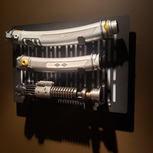 May include: Three silver and black lightsaber props mounted on a black wall mount with a grid pattern. The lightsabers have a yellow accent on the hilt.