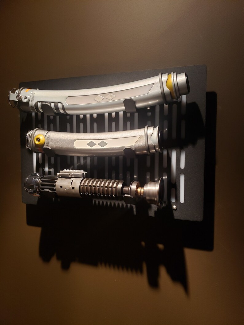 May include: Three silver and black lightsaber props mounted on a black wall mount with a grid pattern. The lightsabers have a yellow accent on the hilt.