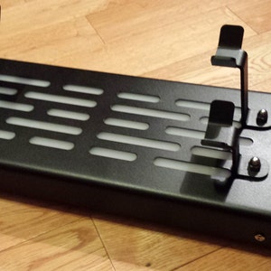 May include: Black metal stand with two metal brackets and a grid pattern. The stand is designed to hold two items, such as controllers or phones.