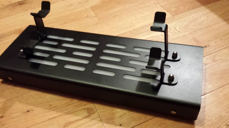 May include: Black metal stand with two metal brackets and a grid pattern. The stand is designed to hold two items, such as controllers or phones.