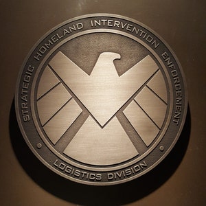 May include: A circular, metallic emblem with the text "STRATEGIC HOMELAND INTERVENTION ENFORCEMENT LOGISTICS DIVISION" encircling a central eagle and shield design. The emblem is a muted silver colour and is set against a dark brown background.