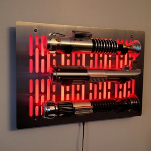 May include: A metal wall mount with a red light behind it, displaying three lightsabers. The lightsabers are silver and black with a red glow.
