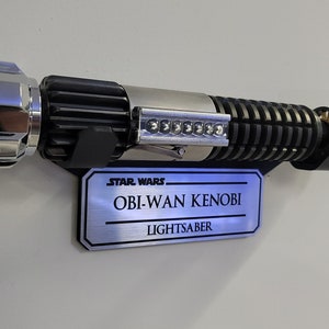 May include: A silver and black Star Wars Obi-Wan Kenobi lightsaber replica with a silver plaque that reads "STAR WARS - OBI-WAN KENOBI - LIGHTSABER".