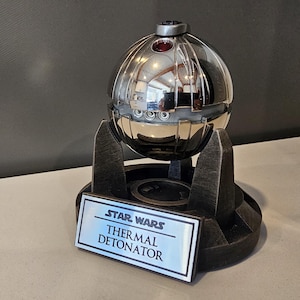 Peut inclure: Une réplique argentée du détonateur thermique Star Wars sur un socle noir avec une plaque argentée indiquant "Star Wars Thermal Detonator".