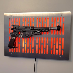 Peut inclure: Un jouet pistolet noir de type blaster monté sur une plaque murale en métal avec un motif de grille de lumière rouge.