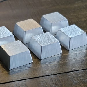 May include: Eight silver-colored metal keycaps with a brushed finish. The keycaps are square and have a slightly raised edge.