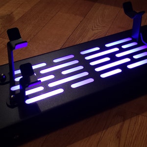May include: A black metal stand with two lightsaber holders and a light-up panel featuring the Star Wars Death Star trench run pattern. The panel is illuminated with a purple light.