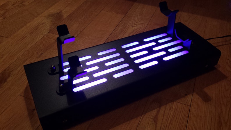 May include: A black metal stand with two lightsaber holders and a light-up panel featuring the Star Wars Death Star trench run pattern. The panel is illuminated with a purple light.