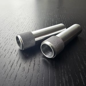 May include: Two cylindrical metal objects with textured grips at one end. The objects are silver in color and appear to be made of stainless steel. They are lying on a dark wooden surface.