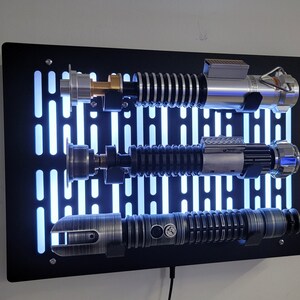 May include: A black wall mount with a Star Wars Death Star design features three lightsabers. The lightsabers are silver, black, and gray with a blue glow.
