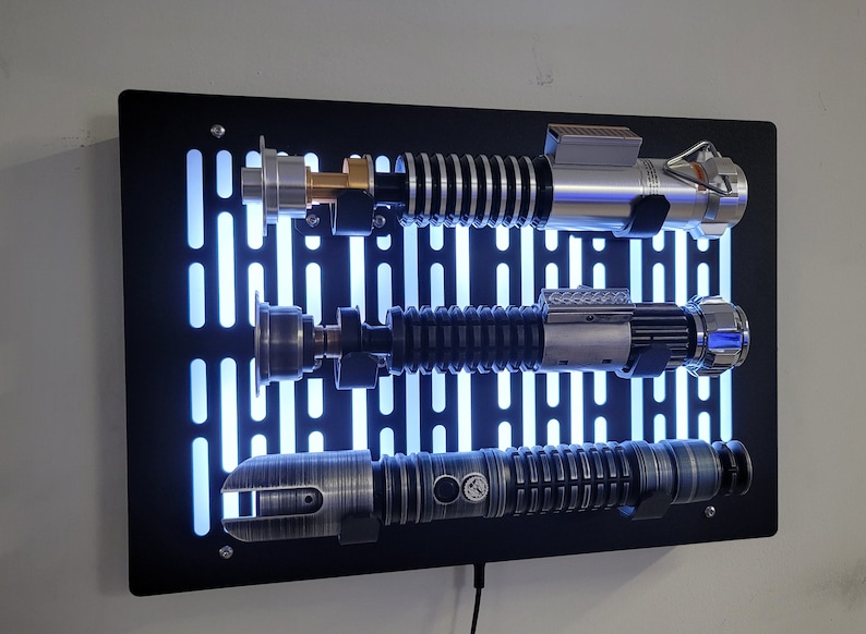 May include: A black wall mount with a Star Wars Death Star design features three lightsabers. The lightsabers are silver, black, and gray with a blue glow.