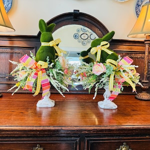May include: Two green bunny figurines with yellow ribbon bows are displayed on white pedestals. Each bunny is surrounded by a floral arrangement with pink, yellow, and white flowers. The arrangements are on a dark wood surface.