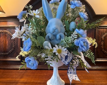 May include: A light blue bunny figurine centerpiece with blue and white flowers and greenery. The bunny is in a white pedestal vase with a blue and white ribbon. The arrangement includes blue roses, daisies, and other floral elements.