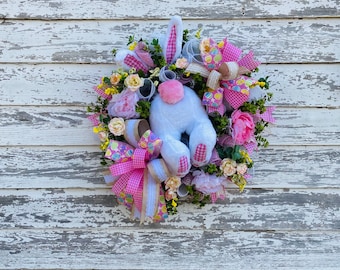 May include: A pink and white Easter bunny wreath with pink and white ribbon, pink flowers, and pastel colored Easter eggs.