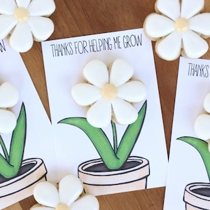 May include: White daisy-shaped cookies with yellow centers are arranged around cards that read "THANKS FOR HELPING ME GROW." The cards feature a drawing of a flower in a pot with green leaves. The cookies and cards are on a wooden surface.
