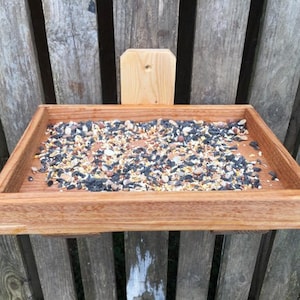 May include: A rectangular wooden bird feeder filled with birdseed. The feeder is made of light brown wood and is mounted on a wooden fence. The birdseed is a mix of seeds and grains. The feeder is designed to attract birds to a garden or outdoor space.