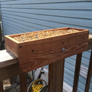 May include: A wooden bird feeder filled with birdseed is mounted on a wooden deck railing. The feeder is made of natural wood with a light brown stain. The feeder is attached to the railing with a metal screw.
