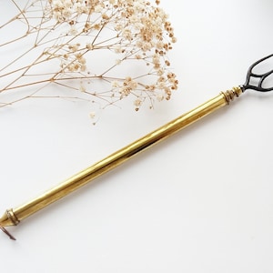 Könnte beinhalten: Ein goldfarbenes Metallwerkzeug mit einem schwarzen, geweihartigen Griff. Das Werkzeug hat ein spitzes Ende und eine kleine Schlaufe am anderen Ende.