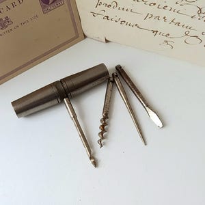 May include: Vintage metal multi-tool with a corkscrew, awl, and other implements. A postcard and handwritten note are in the background. The tool is likely used for opening bottles and other small tasks.