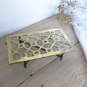 May include: A rectangular brass trivet with a geometric openwork pattern. The trivet has four small legs and is designed to protect surfaces from hot items. The brass has a warm, aged patina.