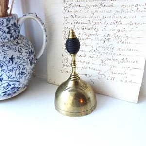 Puede incluir: Una campana de lat&oacute;n vintage con un mango de goma negro. La campana tiene una base redondeada y un mango largo y delgado. La campana est&aacute; sobre una superficie blanca junto a una jarra floral azul y blanca y un trozo de papel envejecido con texto manuscrito.