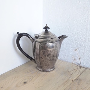 Puede incluir: Una tetera de plata con un asa negra y un diseño decorativo. La tetera tiene un cuerpo redondo y una tapa con un pomo en la parte superior.