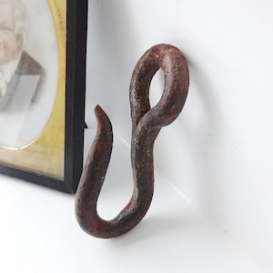 May include: A vintage, rusty metal hook with a curved shape. The hook is a dark brown colour, likely made of iron. It's set against a white background, with a framed portrait visible in the background.