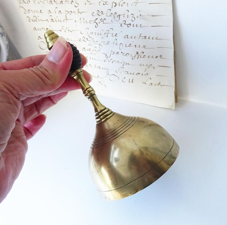 Puede incluir: Una campana de lat&oacute;n vintage con un mango oscuro. La campana tiene una forma redondeada con l&iacute;neas horizontales cerca de la base. El mango tiene un dise&ntilde;o decorativo. Un documento manuscrito est&aacute; en el fondo.