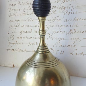 Puede incluir: Campana antigua de lat&oacute;n con un mango de madera negra. La campana tiene una base redondeada con l&iacute;neas grabadas horizontales y un tallo largo. El mango tiene un dise&ntilde;o oscuro y acanalado. La campana es probablemente de principios del siglo XX.