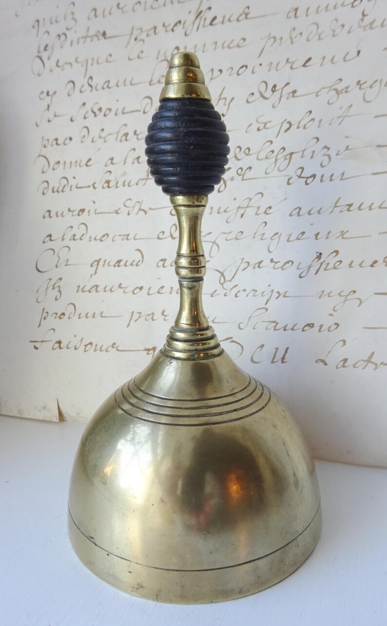 Puede incluir: Campana antigua de lat&oacute;n con un mango de madera negra. La campana tiene una base redondeada con l&iacute;neas grabadas horizontales y un tallo largo. El mango tiene un dise&ntilde;o oscuro y acanalado. La campana es probablemente de principios del siglo XX.