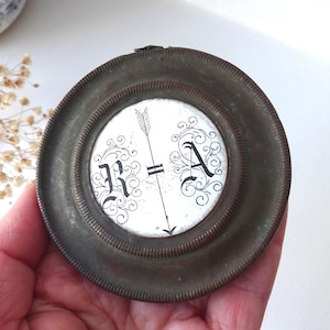 Könnte beinhalten: Ein kleiner, runder, antiker Metallrahmen mit weißem Innenraum. Im Inneren befinden sich die Buchstaben "R" und "A" mit dekorativen Schnörkeln, die von einem Pfeil durchschnitten werden. Der Rahmen hat eine dunkle, gealterte Oberfläche.