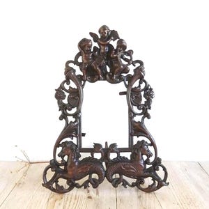May include: Ornate bronze-colored picture frame with cherubs at the top and seahorse-like creatures at the base. The frame has an open center for a photo or artwork, and is made of a detailed, sculpted metal.