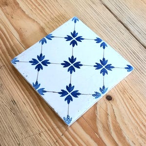 May include: Square white tile with a blue floral pattern. The tile features a repeating design of stylized blue flowers connected by thin lines. The tile is set against a wooden background.
