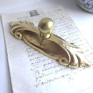 Könnte beinhalten: Verzierte Türgriff aus Messing mit kugelrundem Griff. Die antike Hardware weist ein dekoratives, fließendes Design auf. Der Griff befindet sich vor dem Hintergrund von gealtertem Papier mit handschriftlichem Text, was eine Vintage-Ästhetik suggeriert.