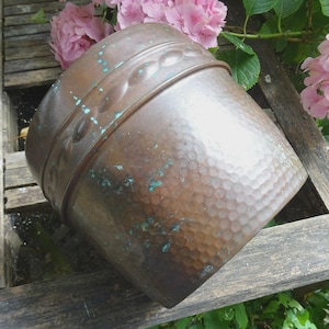 May include: A copper-colored metal planter with a hammered texture and a decorative band near the top. The planter has a square base and a slightly flared top. Patina adds a touch of green to the copper. Pink flowers are in the background.