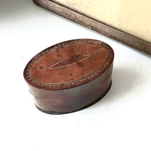 May include: An oval antique leather box, dark brown in colour, with a lighter brown lid. The lid has a central, elongated design and a decorative border. The box is likely for storing small items, such as jewellery or trinkets.