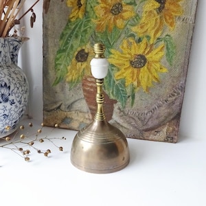 以下が含まれることがあります： 白いセラミックのハンドルが付いたヴィンテージの真鍮のベル。ベルは円錐形のベースと、詳細なリブのデザインが特徴です。ベルはひまわりの絵の前に展示されています。