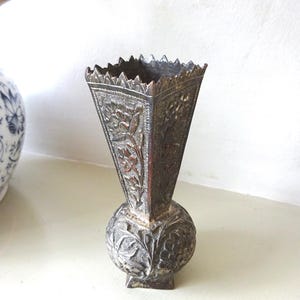 Könnte beinhalten: Eine Vintage-Metallvase mit aufwendigen Blumenschnitzereien. Die Vase hat einen quadratischen Sockel, einen kugelförmigen Mittelteil und eine ausgestellte, dreieckige Oberseite mit dekorativem Rand. Das Metall hat eine dunkle, gealterte Patina.