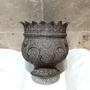 Può includere: Una ciotola antica in metallo scuro con intricati motivi floreali e a mezzaluna. La ciotola ha un bordo decorativo smerlato e una base dettagliata. Il design suggerisce un'influenza mediorientale o asiatica, perfetta per l'arredamento della casa.