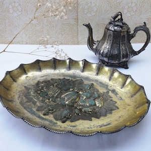 Może przedstawiać: Srebrny czajnik z kwiatowym wzorem stoi na ozdobnym, falowanym, złoconym talerzu do serwowania z wytłoczonym wzorem kwiatowym.