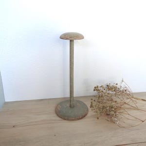 May include: A vintage hat stand with a round base and a tall, slender pole supporting a rounded top. The stand is painted in muted green and brown tones, with visible wear and tear, suggesting an antique aesthetic. The stand is placed on a wooden surface.